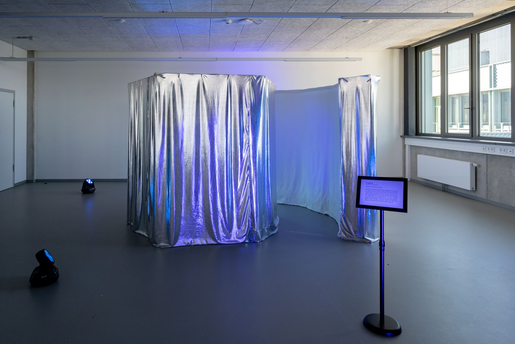 Die Installation: eine mit silbernem Lamé verhüllte Kabine in einem hellen Raum, blau-violett beleuchtet, daneben ein Display mit dem Ausstellungstext.
